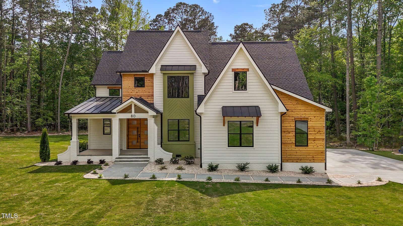 Two-story house with white and wood siding, black roof, and modern architectural features. There is a covered front porch, large windows, and a driveway to the right, surrounded by lawn and trees.