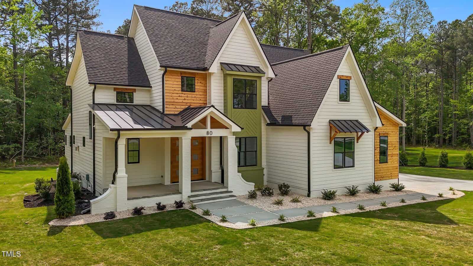 Two-story modern farmhouse with white siding, wood accents, black window frames, covered front porch, and landscaped yard, set against a backdrop of trees and greenery.