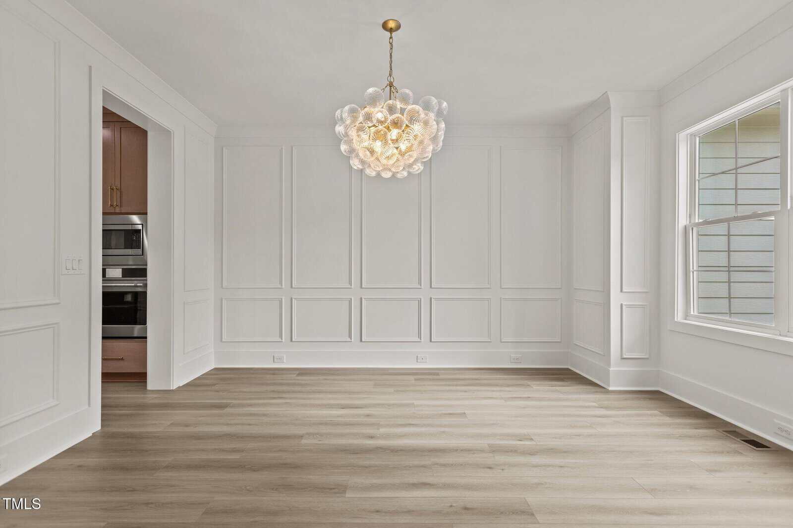 A dining room with white paneled walls, light wood flooring, a bubble glass chandelier, and a large window. An entryway leads to a kitchen with built-in appliances visible. The space is unfurnished and well-lit.