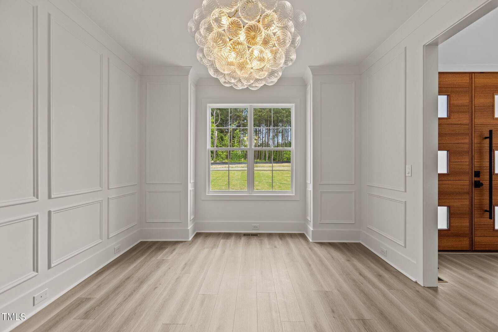 A bright, empty room with light wood flooring, white paneled walls, a large window showing green grass and trees outside, and a round, modern chandelier hanging from the ceiling.