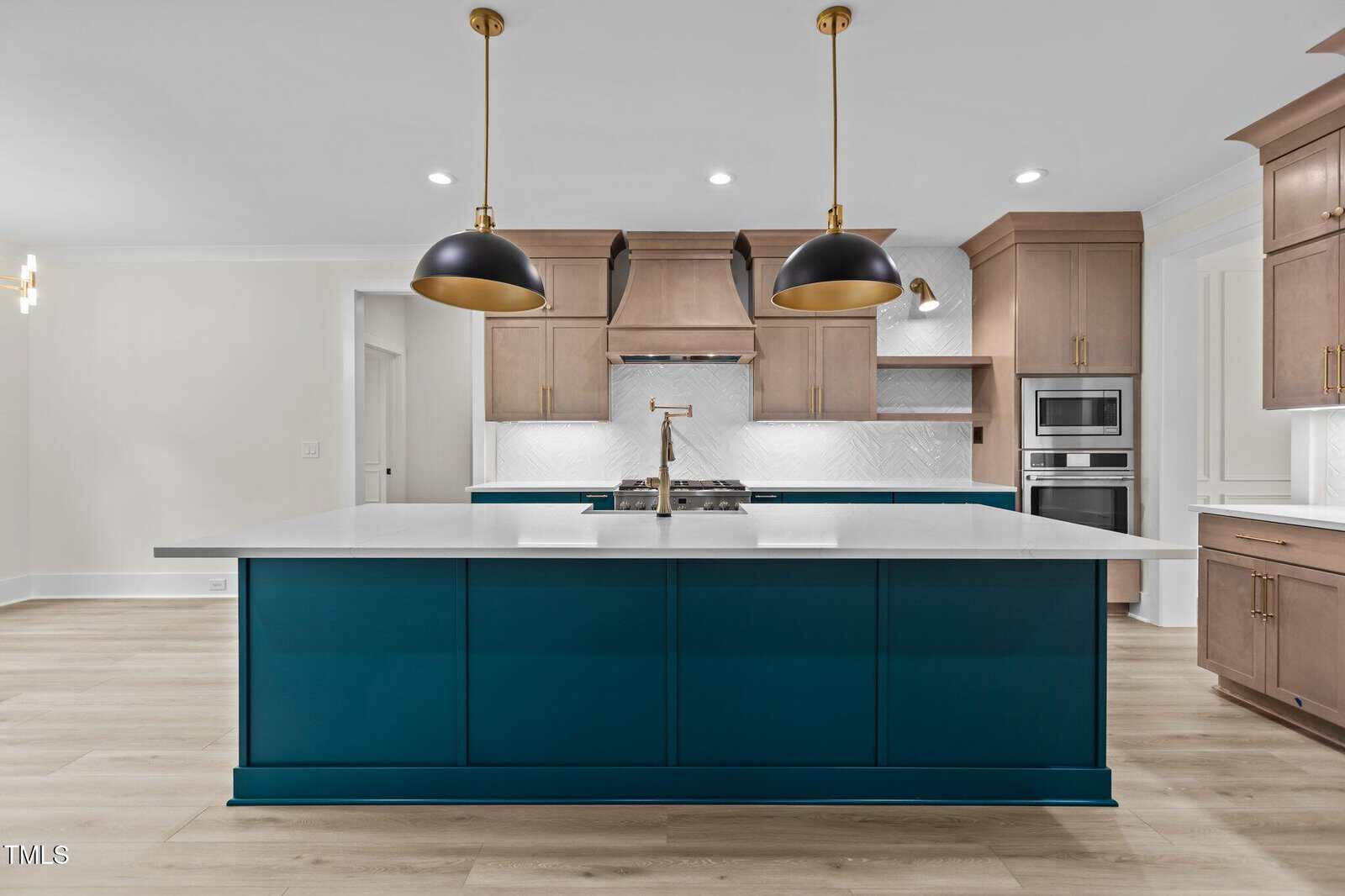 Modern kitchen with a large blue island, white countertops, gold faucet, two black pendant lights, light wood cabinets, and built-in stainless steel oven and microwave. Light wood floors and white walls complete the space.