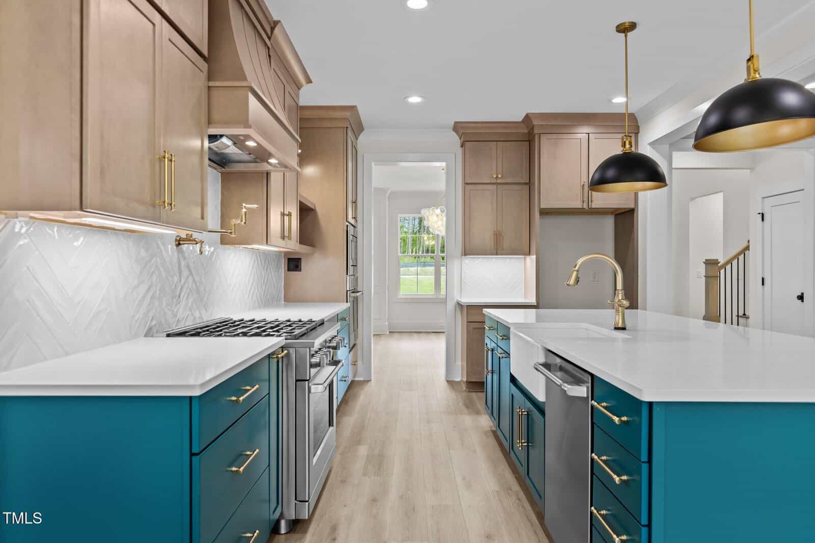 Modern kitchen with wood cabinets, blue lower cabinets, white countertops, stainless steel appliances, gold hardware, a farmhouse sink, two pendant lights, and light wood floors. A dining area is visible through a doorway.