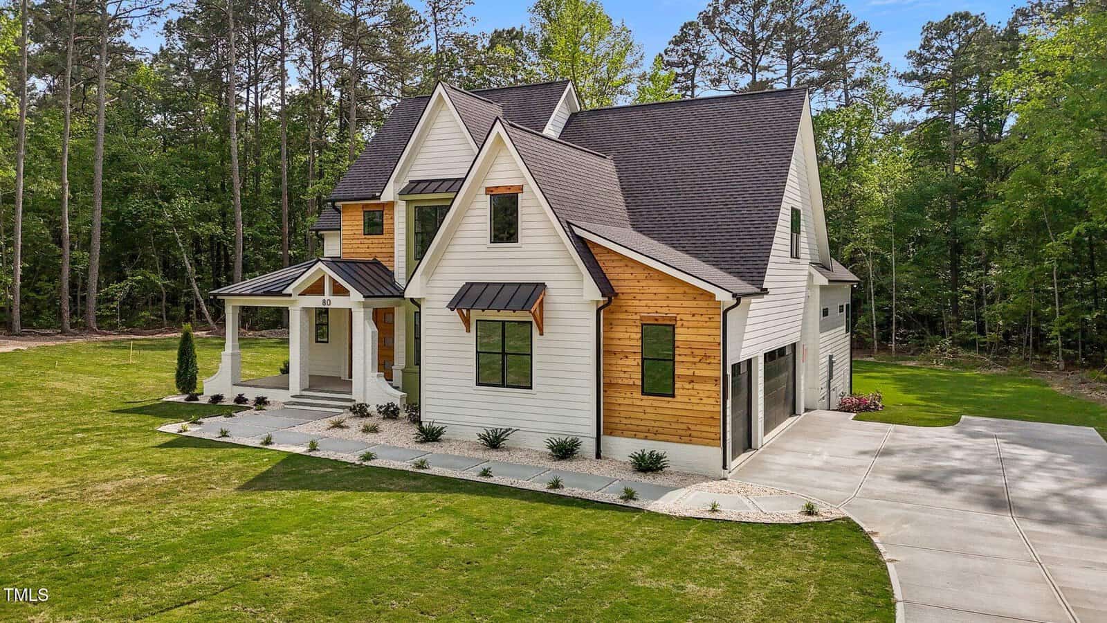 A modern two-story house with white siding and wood accents, black-framed windows, and a covered front porch. The home sits on a large, well-manicured lawn surrounded by trees with a concrete driveway.