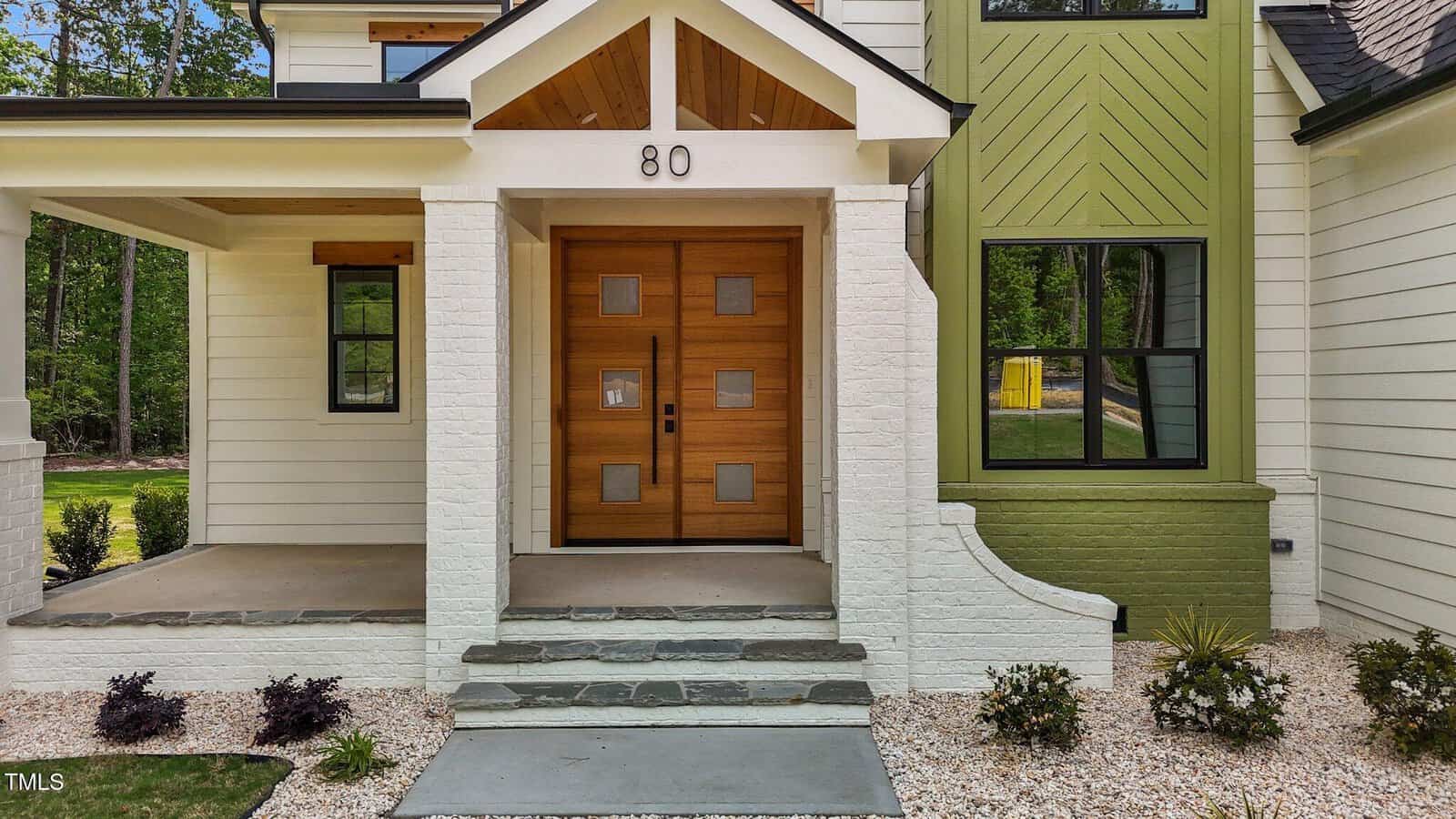 Contemporary house entrance with double wooden front doors featuring square glass panels, white exterior walls, green accent wall, small front porch, and minimal landscaping. The number 80 is displayed above the door.
