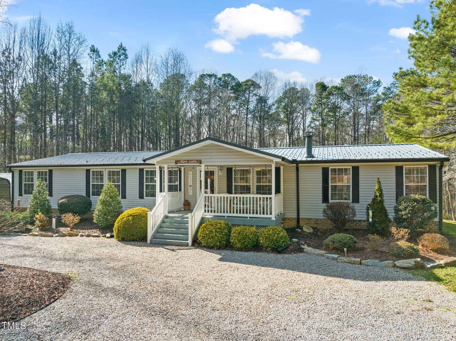 Located at 3041 Jacksontown Road in Manson, this charming single-story house boasts light gray siding and dark shutters. It features a metal roof, a covered porch with steps, and is embraced by a gravel driveway, manicured shrubs, and trees beneath a partly cloudy sky.
