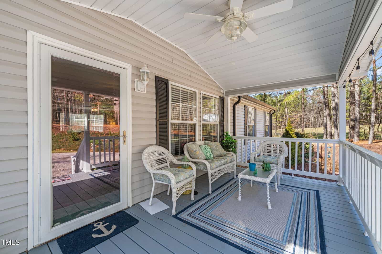 A covered porch features wicker furniture with cushions, a small table, and a ceiling fan. The area is enclosed by a white railing and overlooks a yard with trees. The porch has gray flooring and a welcome mat near the glass door.