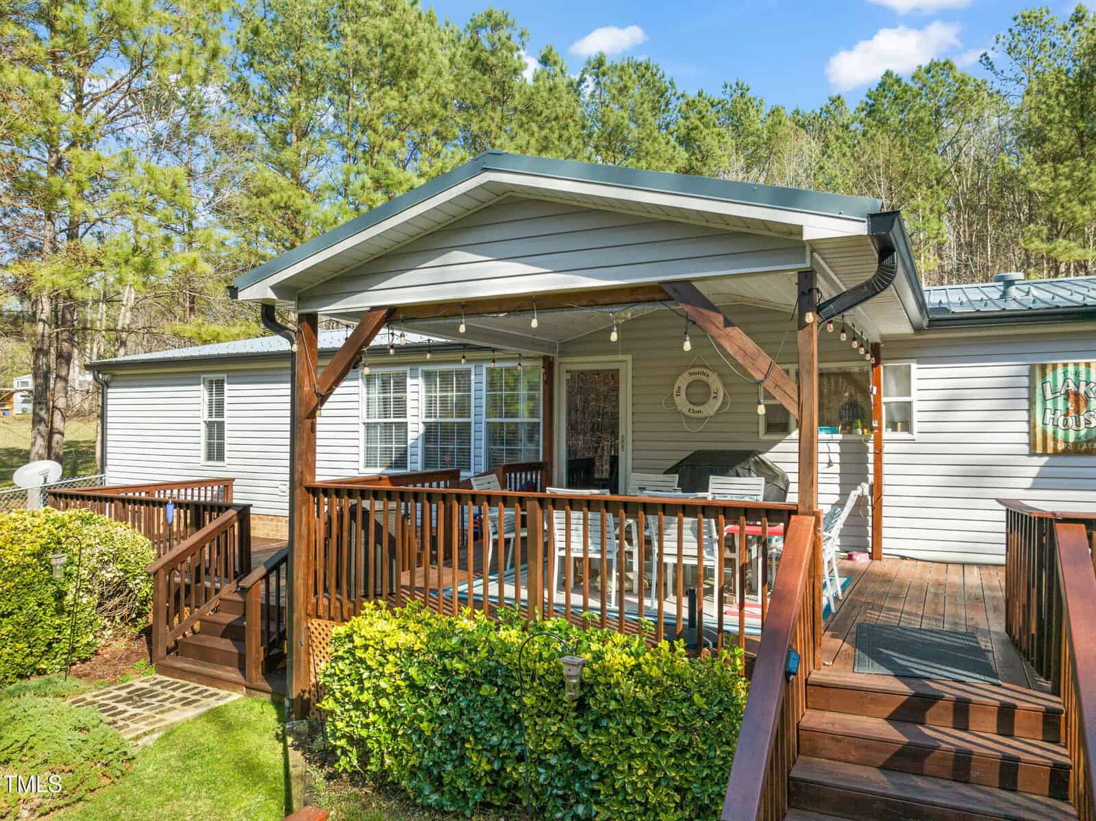 A wooden deck with a covered porch attached to a gray house. The deck has railings and stairs leading down to a green lawn. There are string lights hanging from the porch roof. Trees are visible in the background under a clear blue sky.