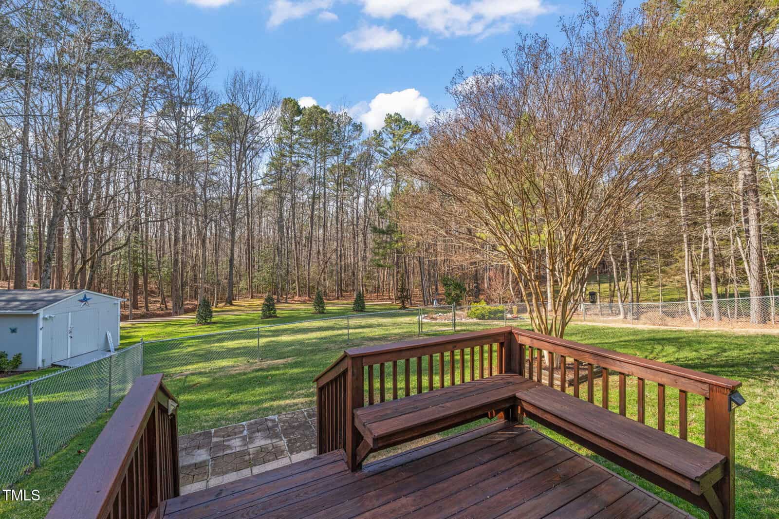 View from a wooden deck overlooking a spacious backyard. The yard features a mix of grass, trees, and shrubs, bordered by a wire fence. In the distance, a shed is visible on the left, and tall trees line the background under a partly cloudy sky.