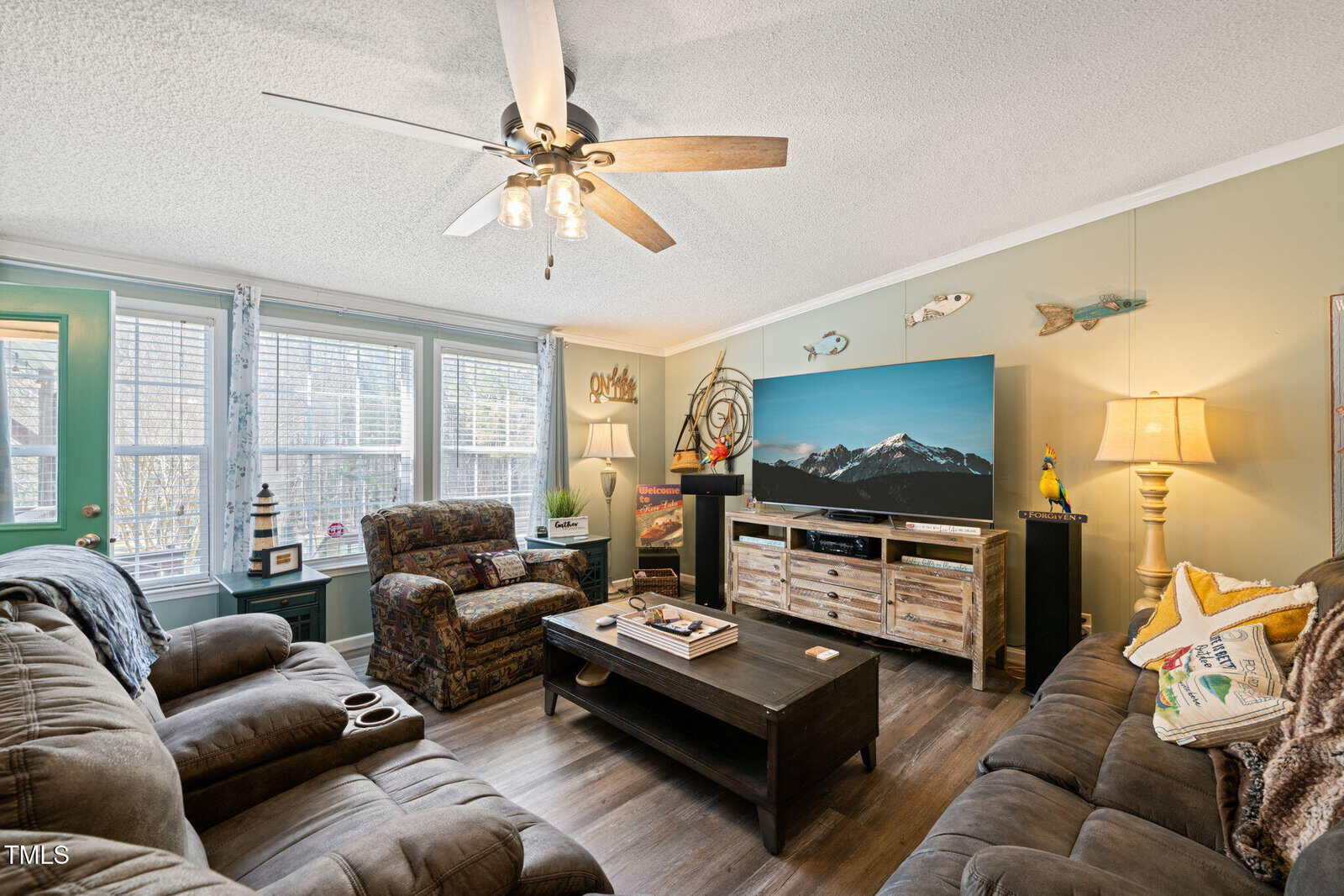 A cozy living room with two couches and an armchair facing a flat-screen TV. The room features a ceiling fan, large windows with natural light, a wooden coffee table, and wall decor including a fish collection and a "Home" sign.