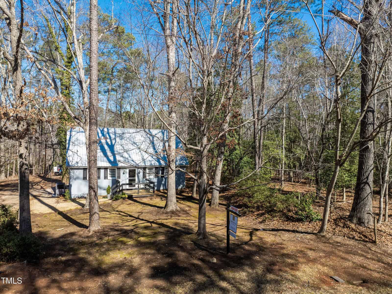 A small white house with a metal roof sits among tall, leafless trees on a sunny day. A dirt driveway leads to the house, and a sign is posted in front of the property. The surrounding area is wooded.