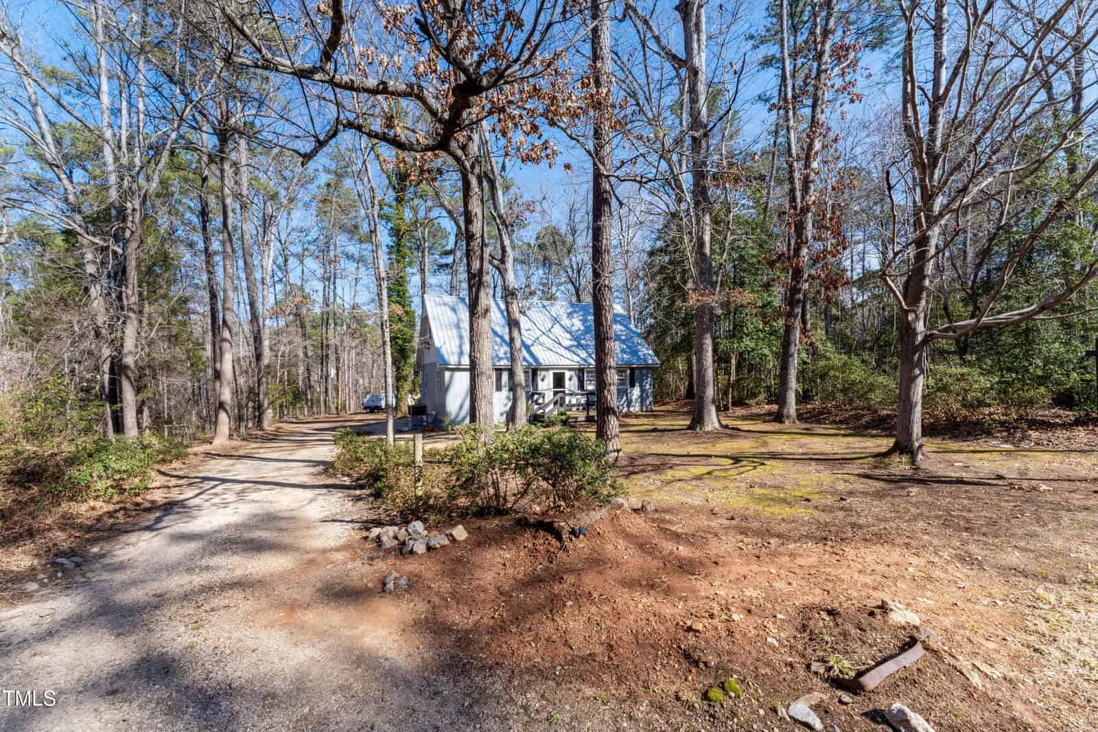 A gravel driveway leads through a wooded area with tall trees to a small white house with a blue metal roof, surrounded by scattered leaves and patches of sunlight.