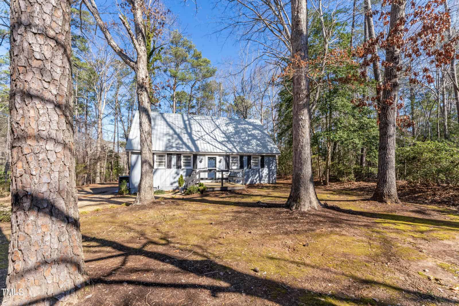 A small white house with a gray roof sits among tall trees on a sunny day. The yard is mostly dirt and fallen leaves with patches of moss, and there is a wooden ramp leading to the front door.