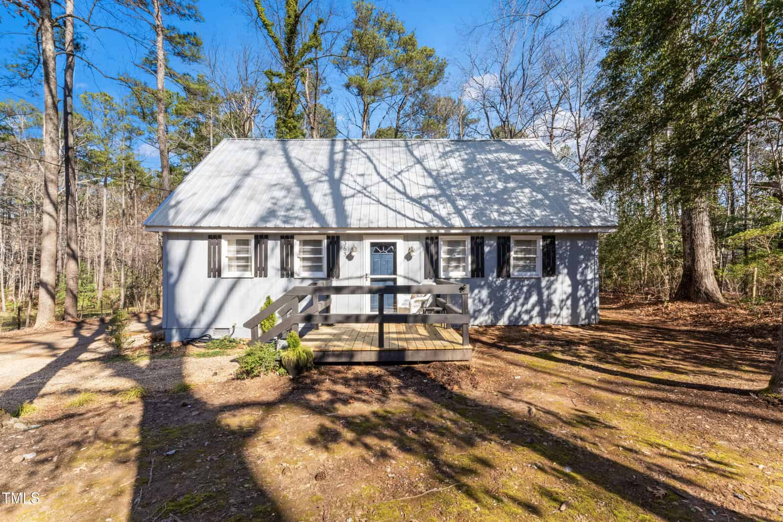 A light blue, two-story house with a steep roof, white shutters, and front porch sits surrounded by tall trees, with a bare dirt and leaf-covered yard in bright sunlight.