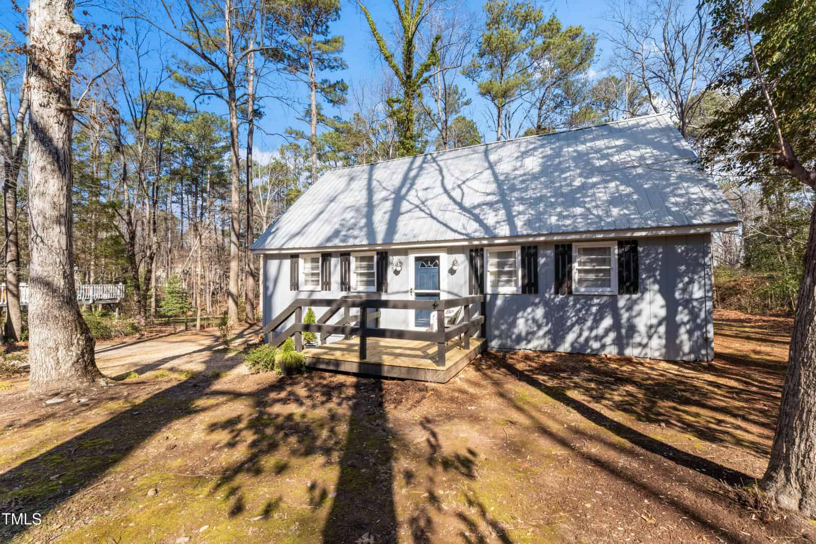 A gray house with a metal roof and a small front porch sits among tall trees on a sunny day. The yard is mostly bare dirt with some plants near the porch. Shadows from the trees cover parts of the house and ground.