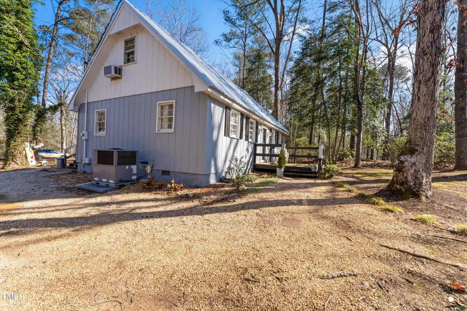 A gray A-frame house with white trim sits in a wooded area. The house has several windows, a small deck with steps, gravel driveway, and an outdoor air conditioning unit. Sunlight filters through the trees.