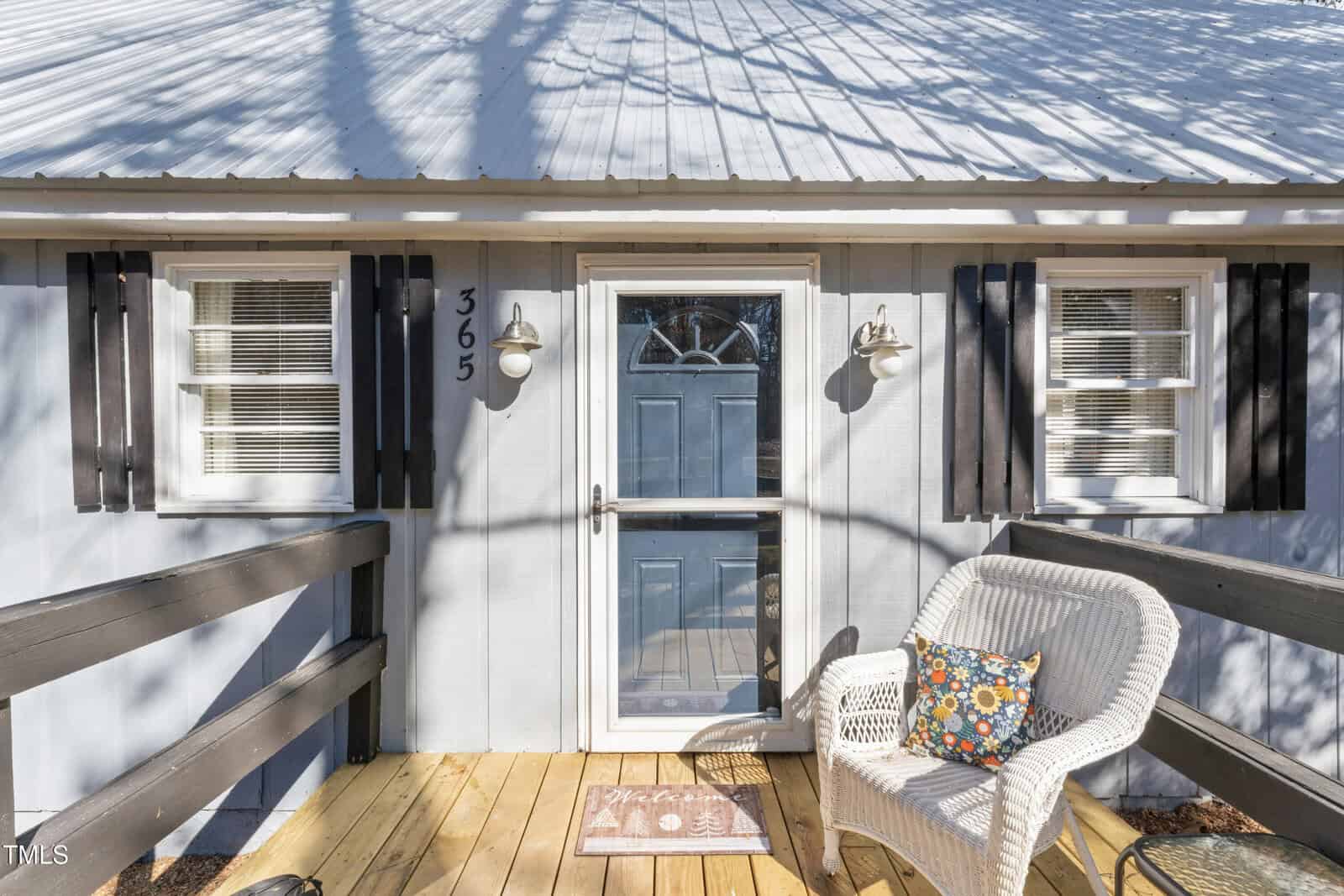 Front entry of a house with a blue door, two windows with shutters, a white wicker chair with a cushion, a patterned doormat, and wooden porch with black railing. House number 365 is visible next to the door.