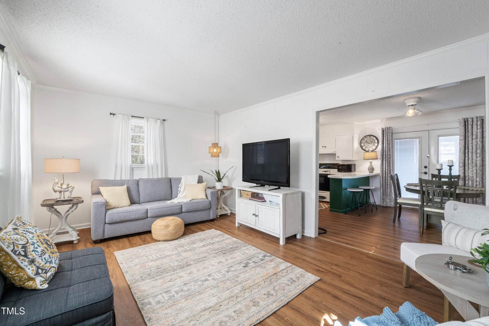 Bright living room with wood floors, gray sofas, a TV on a white stand, floor lamp, neutral rug, and a small table. Kitchen and dining area are visible in the background through an open doorway.