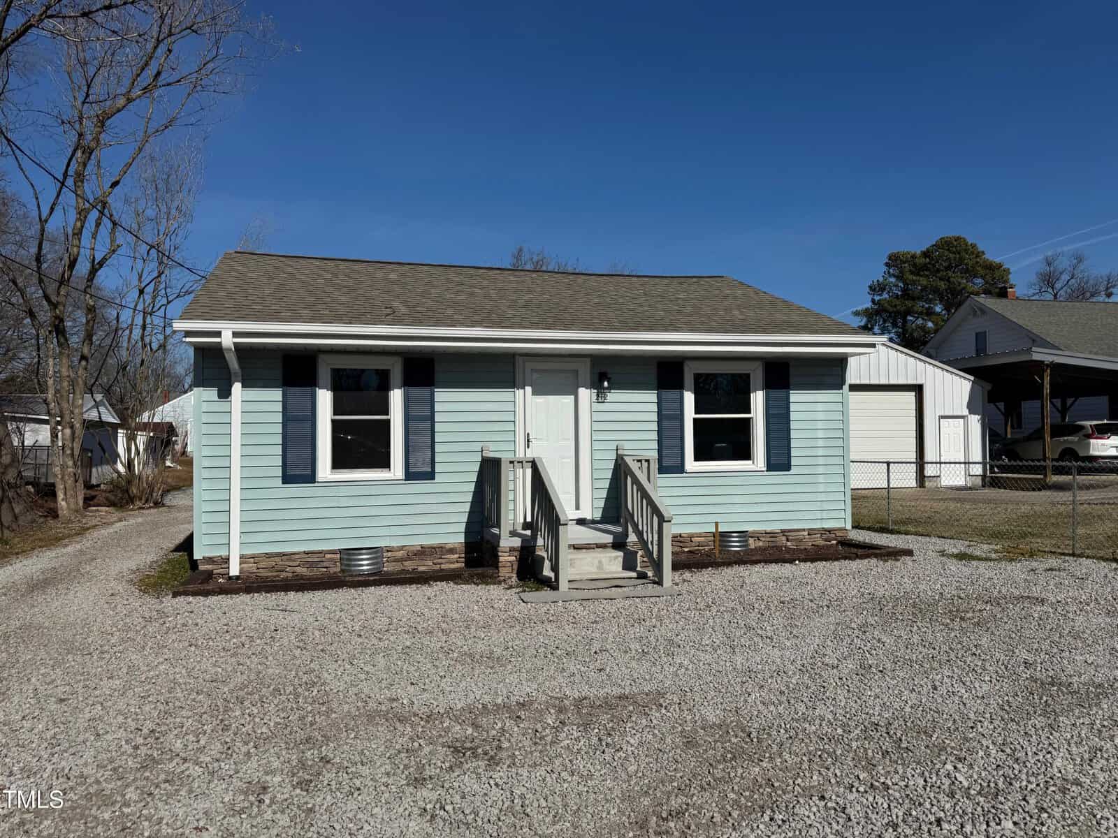 A small light blue house at 212 Craig Avenue features a single-story structure with two windows and a central front door, accessed by wooden steps. The gravel yard is complemented by a detached garage to the right and a few trees gracing the background in Henderson.