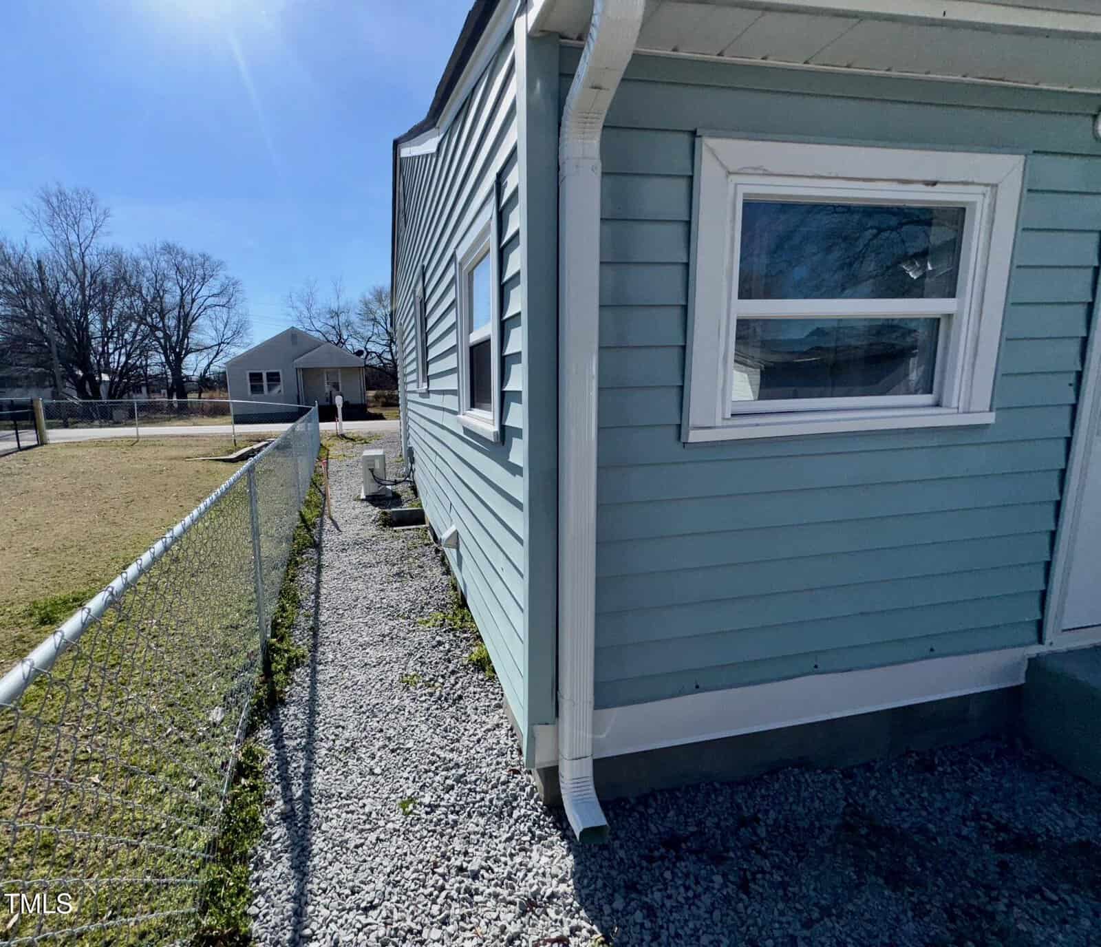 The image shows the side of a light blue house with a gravel path alongside it. A chain-link fence runs parallel to the house. There's a window in view, and trees are visible in the background against a clear blue sky.