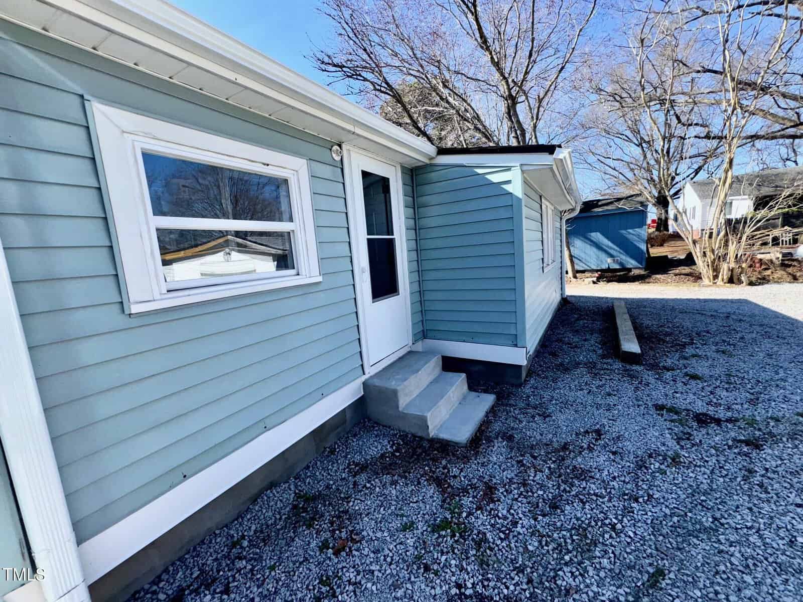 Side view of a light blue house with a white door and a small set of concrete steps. The exterior features horizontal siding. The ground is covered with gravel, and there are leafless trees and another building in the background.