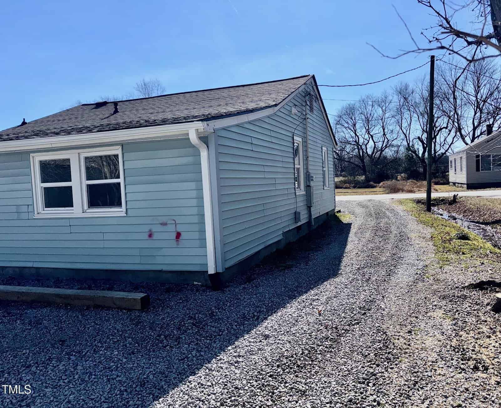 A small, light blue house with a shingled roof is situated on a gravel driveway. There are two windows on the side and graffiti on the wall. In the background, leafless trees and another house are visible under a clear blue sky.
