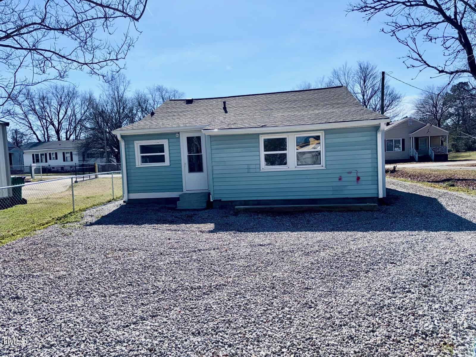 A small blue house with a light gray roof is pictured. It has a few windows and a single door with steps leading up to it. The front yard is covered with gravel and there are leafless trees and other houses in the background.