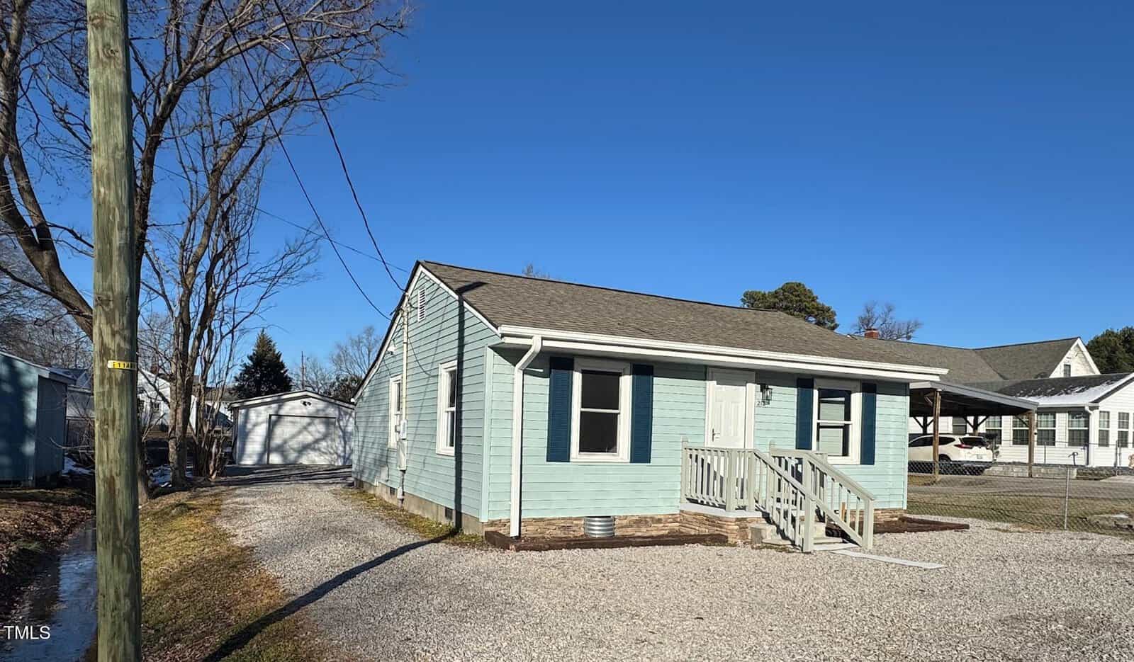 A small, light blue house with a gray roof and white trim. There is a gravel driveway leading to a detached garage in the background. The yard is bordered by leafless trees and the sky is clear and blue.