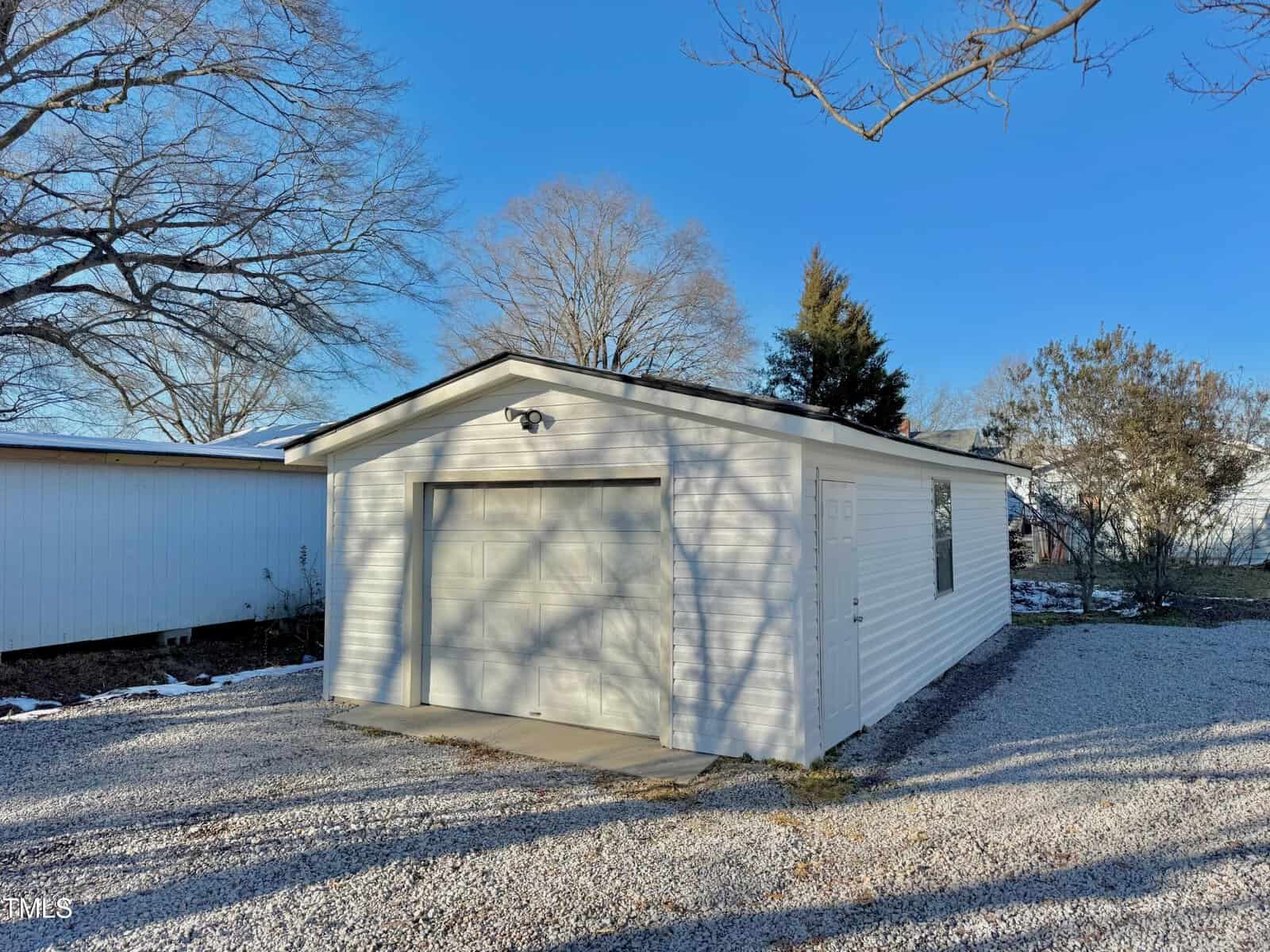 A small white garage with a closed door stands on a gravel driveway. The sky is clear and blue, and trees without leaves surround the structure. The sun casts shadows on the garage and the ground.