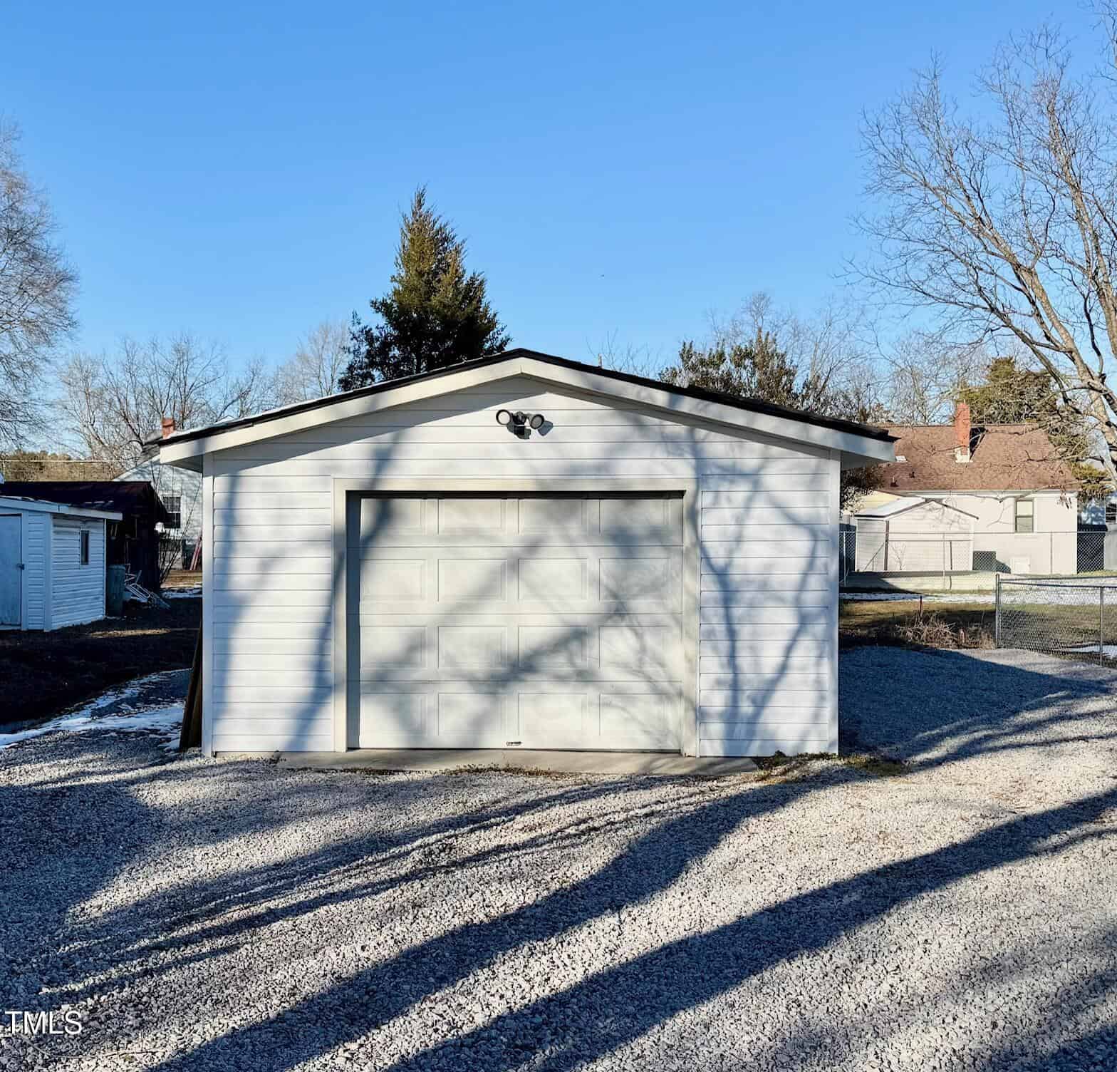 A small, detached garage with a white exterior and a closed garage door is situated on a gravel driveway. Shadows from nearby trees are visible on the building. The sky above is clear and blue, with some residential buildings in the background.