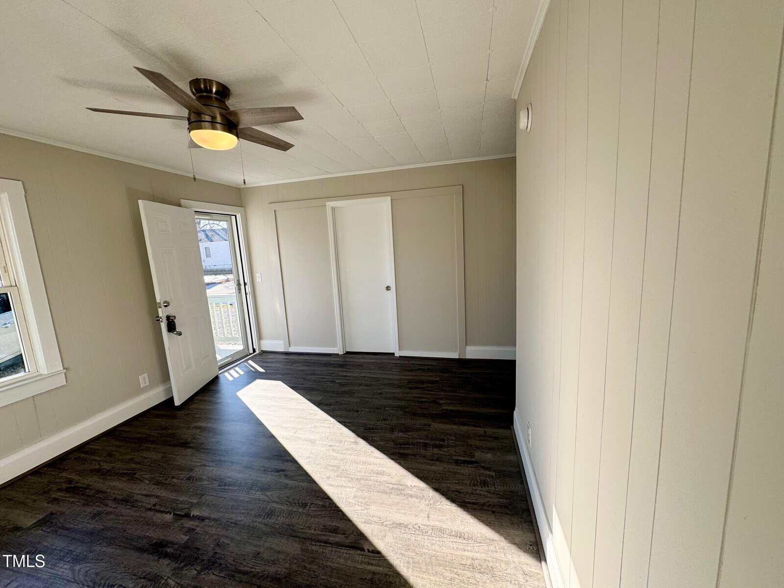 Empty room with light beige walls and dark hardwood floors. The ceiling has a fan with a light. Sunlight enters through a window next to a white door. Built-in closets with sliding doors line one wall.