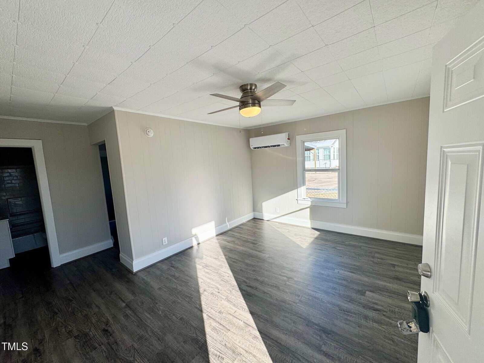 A vacant room with light gray walls and dark wood flooring. The ceiling has a fan mounted in the center. There is a window letting in natural light and a wall-mounted air conditioning unit. An open doorway is visible to the left.