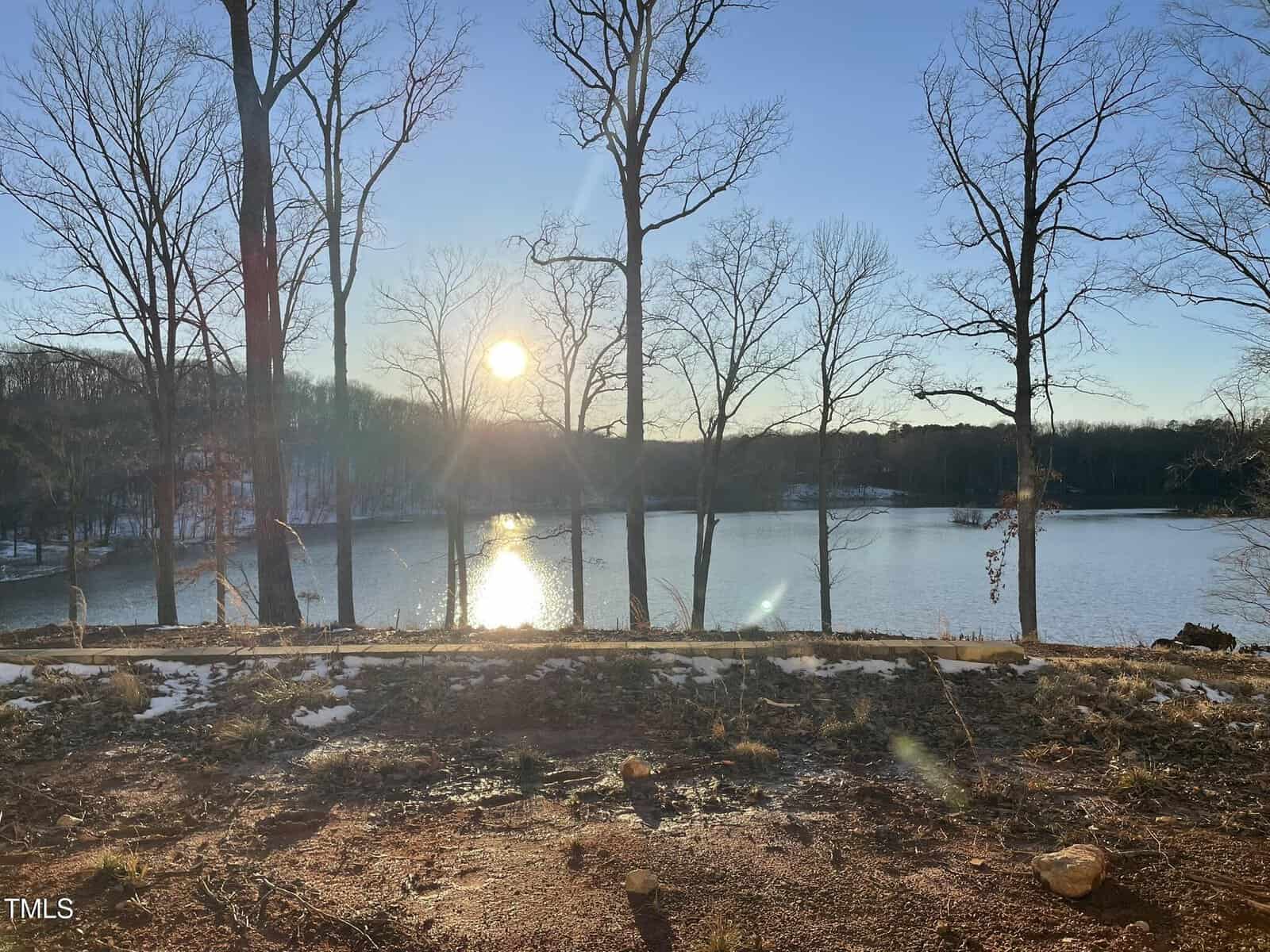 Sunset view over a calm lake with bare trees along the shoreline. The sun reflects on the water's surface, with a clear sky and traces of snow on the ground in the foreground.