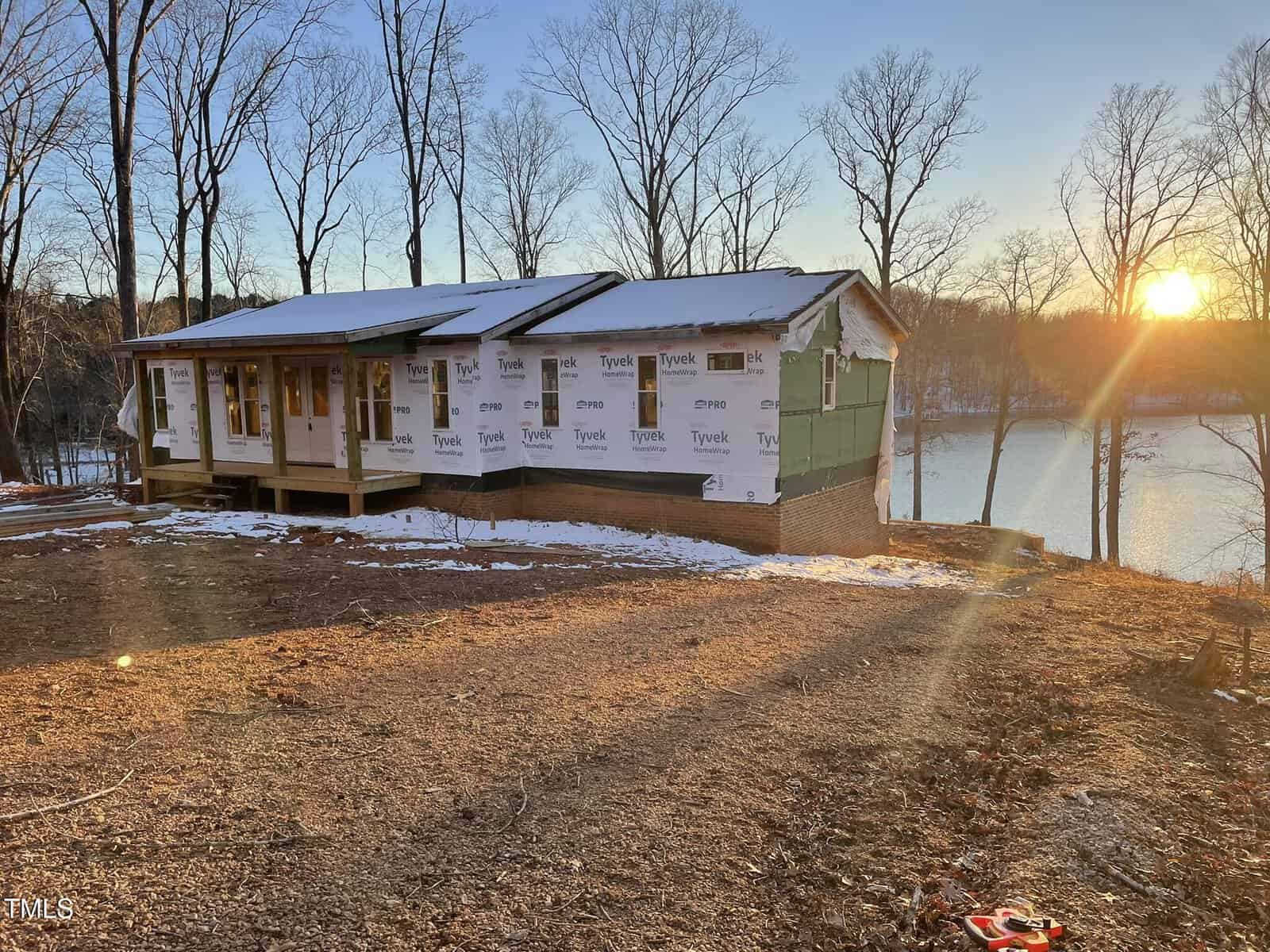 A house under construction with partial walls cloaked in Tyvek wrap stands on a clearing by a lake at 1689 Peninsula Lane. Nearby, leafless trees frame the scene as the setting sun reflects off the water, casting a warm glow across Bullock's property.