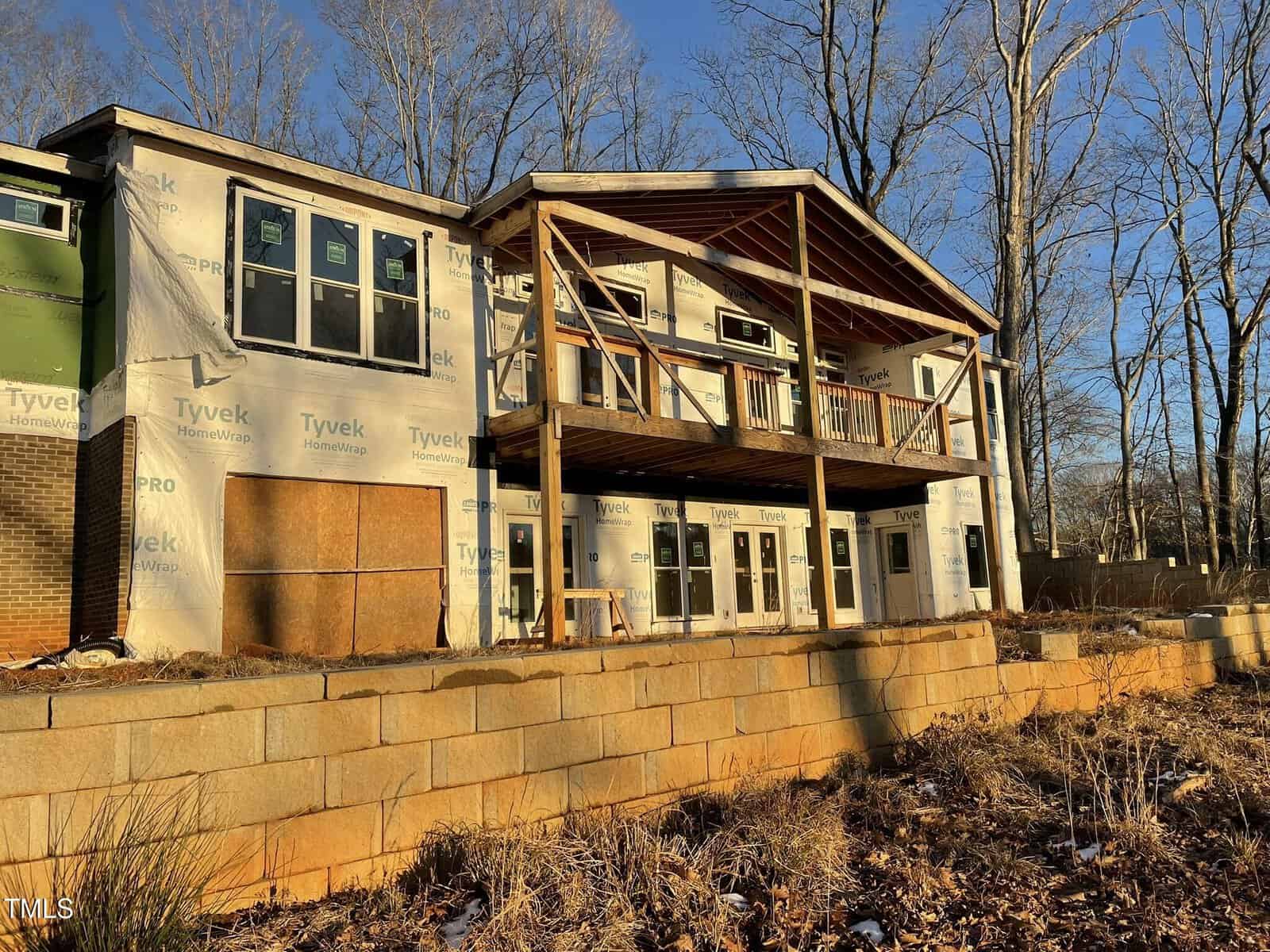 Partially constructed house with exposed wooden beams and a wraparound deck. The exterior walls are covered with Tyvek insulation. Leafless trees and a stone retaining wall are in the background, and the sun casts long shadows.