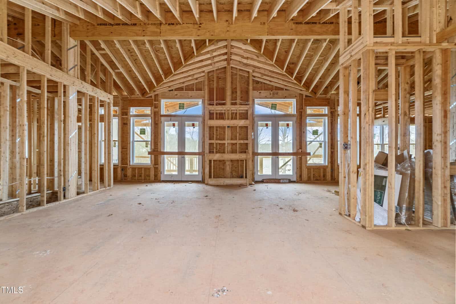 Unfinished interior of a wooden-framed house under construction. Visible wall studs, ceiling beams, and floor. Large windows and doors at the far end, providing natural light. Sparse construction materials are scattered on the floor.