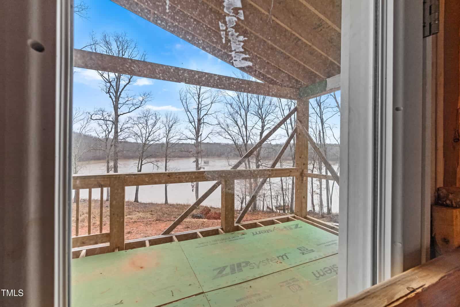 View through a window showing a partially constructed wooden porch facing a lake. The structure has a roof but no railing. Leafless trees stand between the porch and the water under a blue sky. Construction materials are scattered on the porch floor.