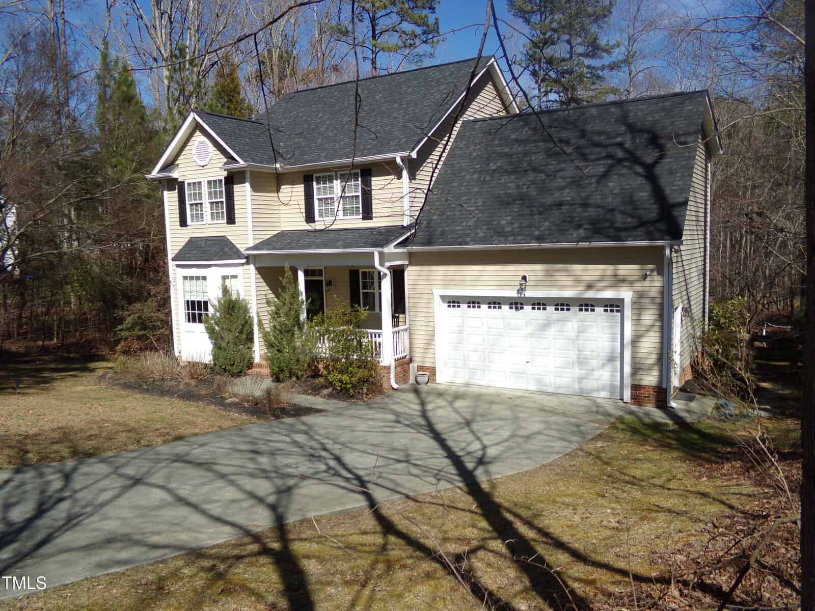 Nestled at 1226 Woodland Church Road in Wake Forest, this two-story beige house boasts a dark roof and white trim. It features a charming covered porch, an attached two-car garage, and a concrete driveway. Small shrubs adorn the front yard, while trees surround the property against a clear blue sky backdrop.