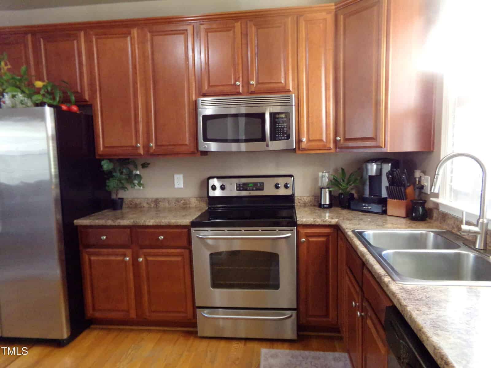 A kitchen featuring wooden cabinets, a stainless steel fridge, and an oven with a microwave above it. The countertop holds a blender and a knife block. A double sink is visible to the right, and a small plant is placed near the corner.