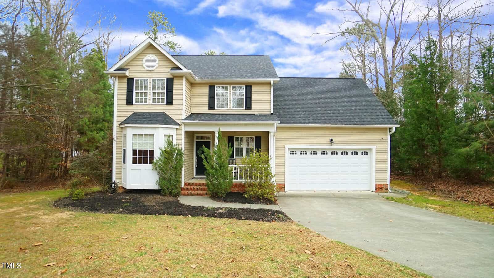A two-story beige house with white trim and a black roof sits with an attached two-car garage. The house is surrounded by trees and has a small front lawn with a concrete driveway leading to the garage. The sky is partly cloudy.