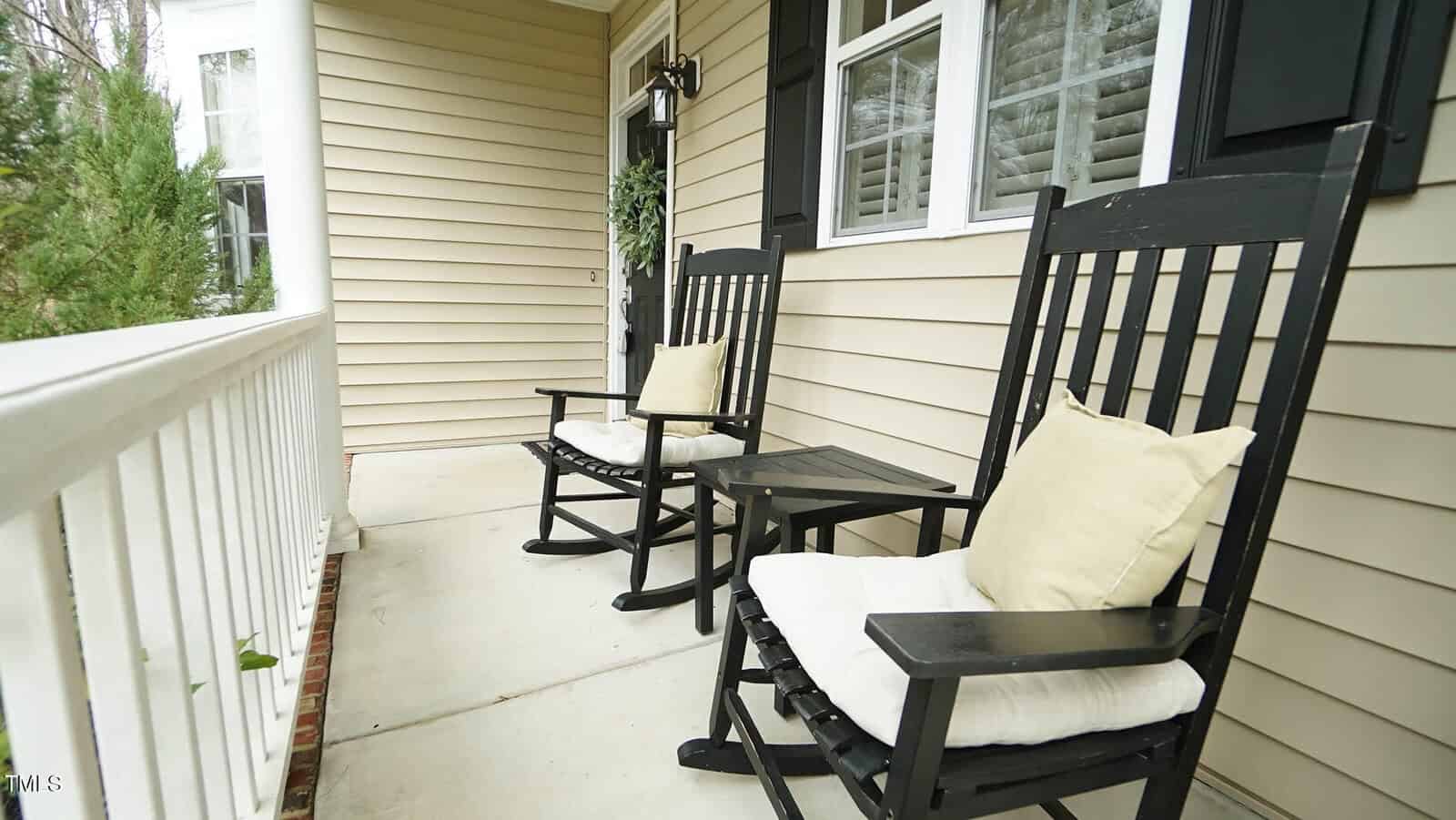 Two black wooden rocking chairs with beige cushions are on a porch with light-colored siding and white railing. A small matching side table is positioned between the chairs. A window with white shutters is on the right side of the wall.