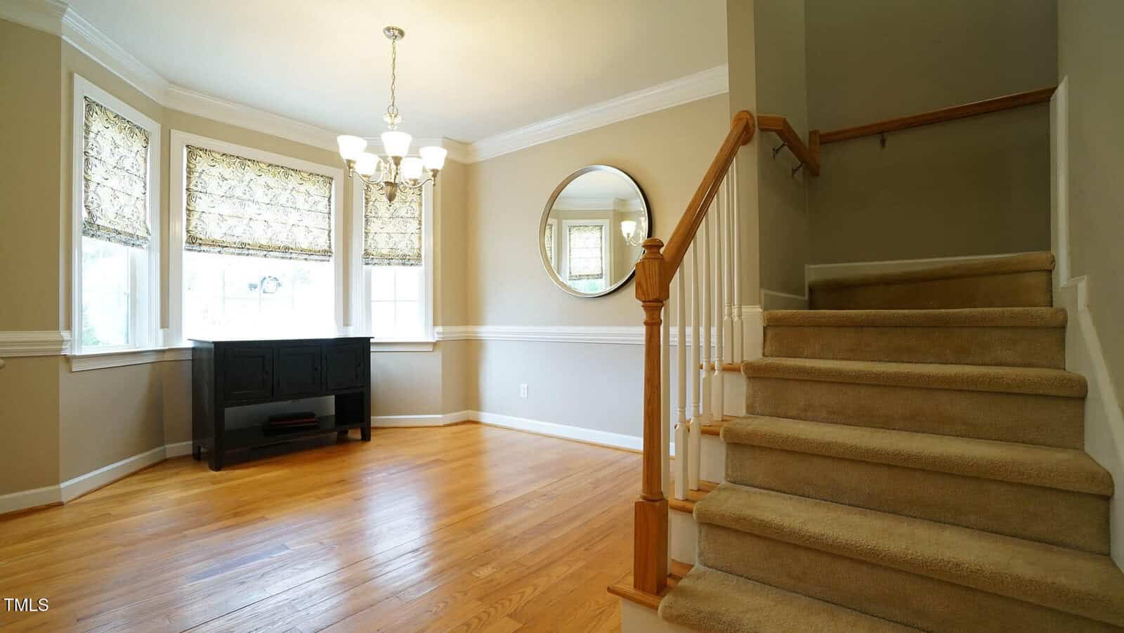 A room with wooden floors, a bay window with patterned curtains, and a black cabinet. A staircase with beige carpet is on the right. A round mirror hangs on the wall above wainscoting, and a chandelier is centered on the ceiling.