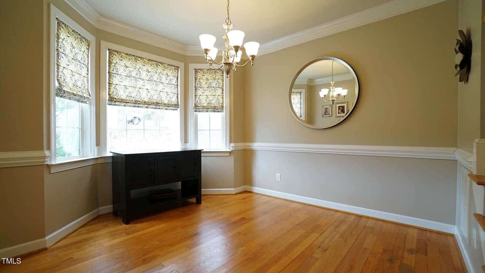 A dining room with hardwood floors, beige walls, and white trim. A round mirror hangs on the right wall. The left side features three windows with patterned shades, and a dark wooden console table is positioned underneath them. A chandelier is centered above.