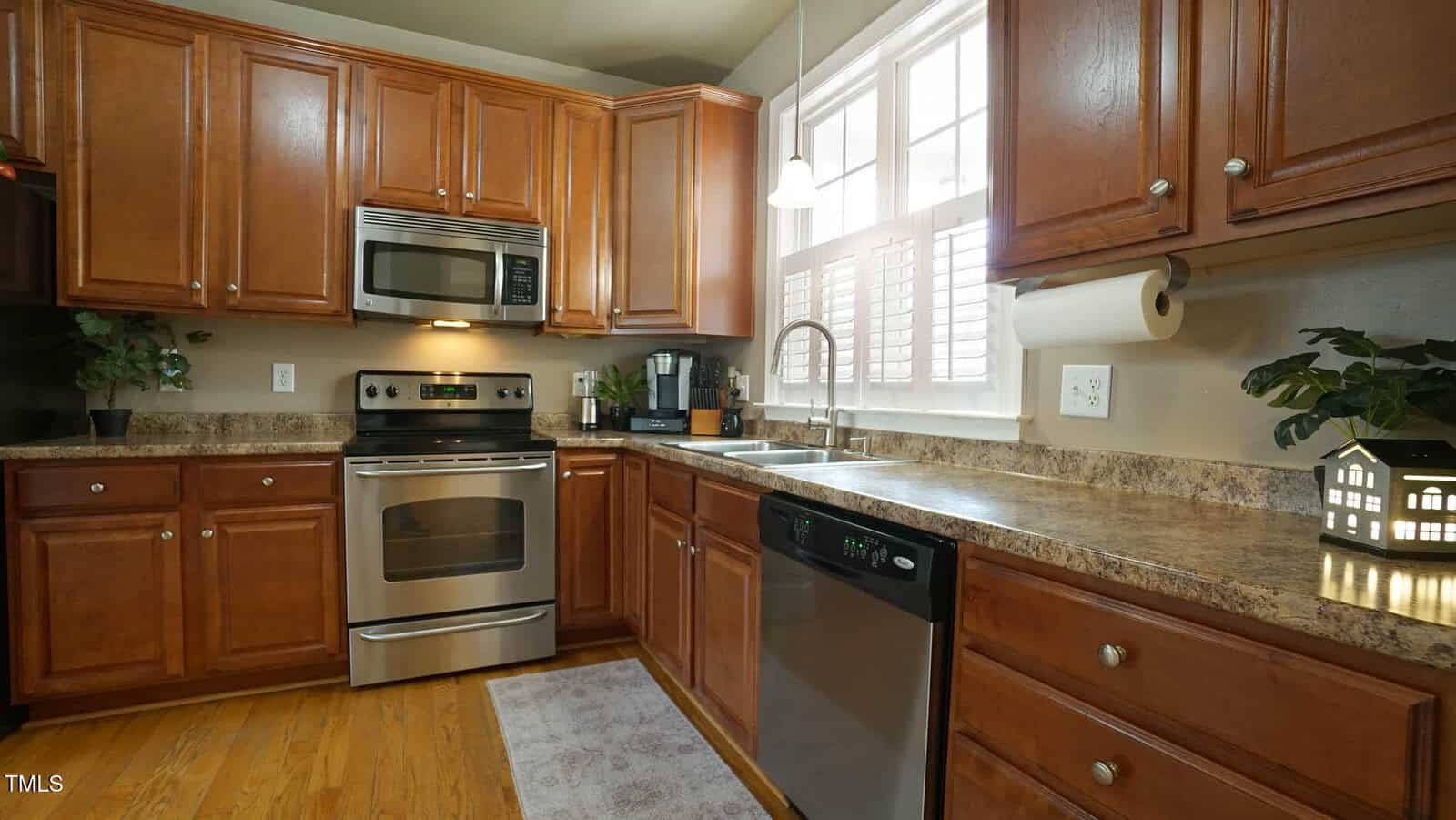 A kitchen with wooden cabinets and a granite countertop. It features a stainless steel oven, microwave, and dishwasher. A window with shutters is above the sink. Various small plants and kitchen items are on the counter. Wood flooring is visible.