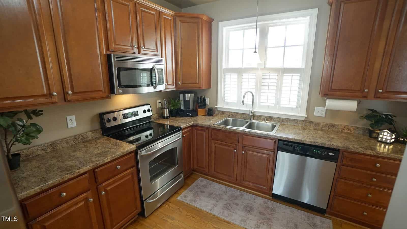A kitchen with wooden cabinetry, stainless steel appliances including an oven and dishwasher, and a double sink under a window. The countertops have a granite-like appearance, and there are small plants and a paper towel holder as decor.