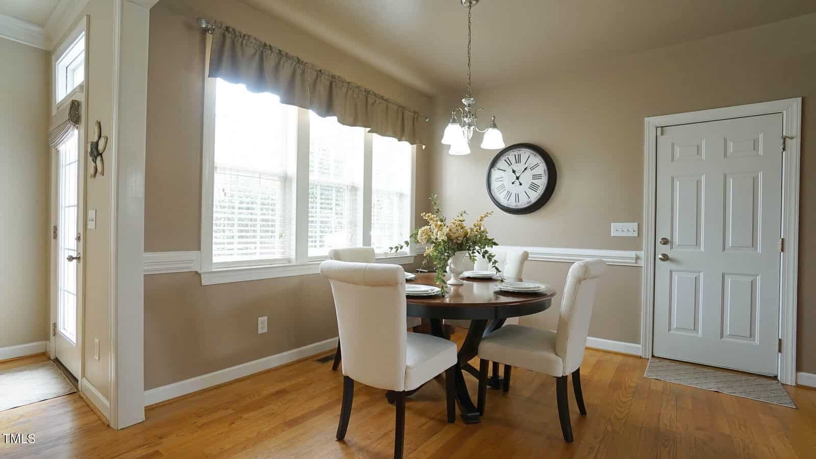A dining area with a round wooden table and four cushioned chairs. A flower centerpiece sits on the table. A large wall clock is mounted above a white door. Windows with blinds and curtains let in natural light. The floor is hardwood.