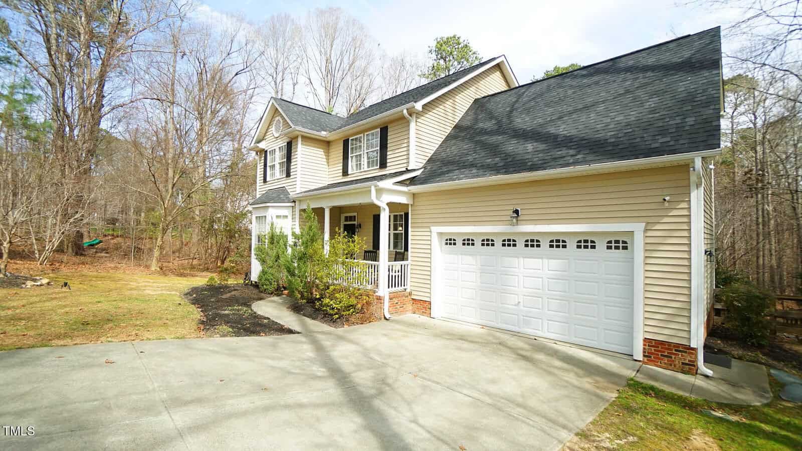 A two-story house with beige siding and a dark gray roof is shown. It features a front porch with white railings, a two-car garage with white doors, and a driveway. The house is surrounded by a grassy yard and trees in the background.