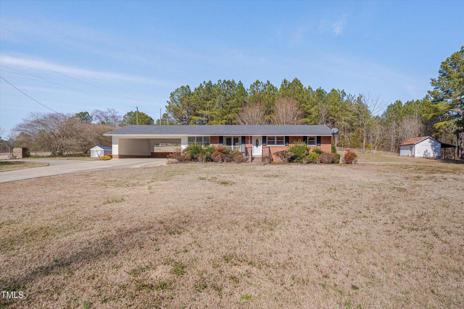A single-story brick house with a dark roof and attached carport sits in the center of a large, dry front yard, bordered by shrubs and surrounded by trees under a clear blue sky.