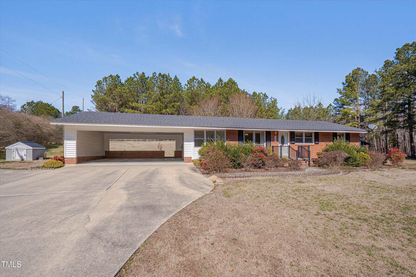 Single-story ranch-style house with a brick and white exterior, attached carport, and a curved driveway. The yard is partially grassy with some shrubs and trees, and a small shed is visible in the background.