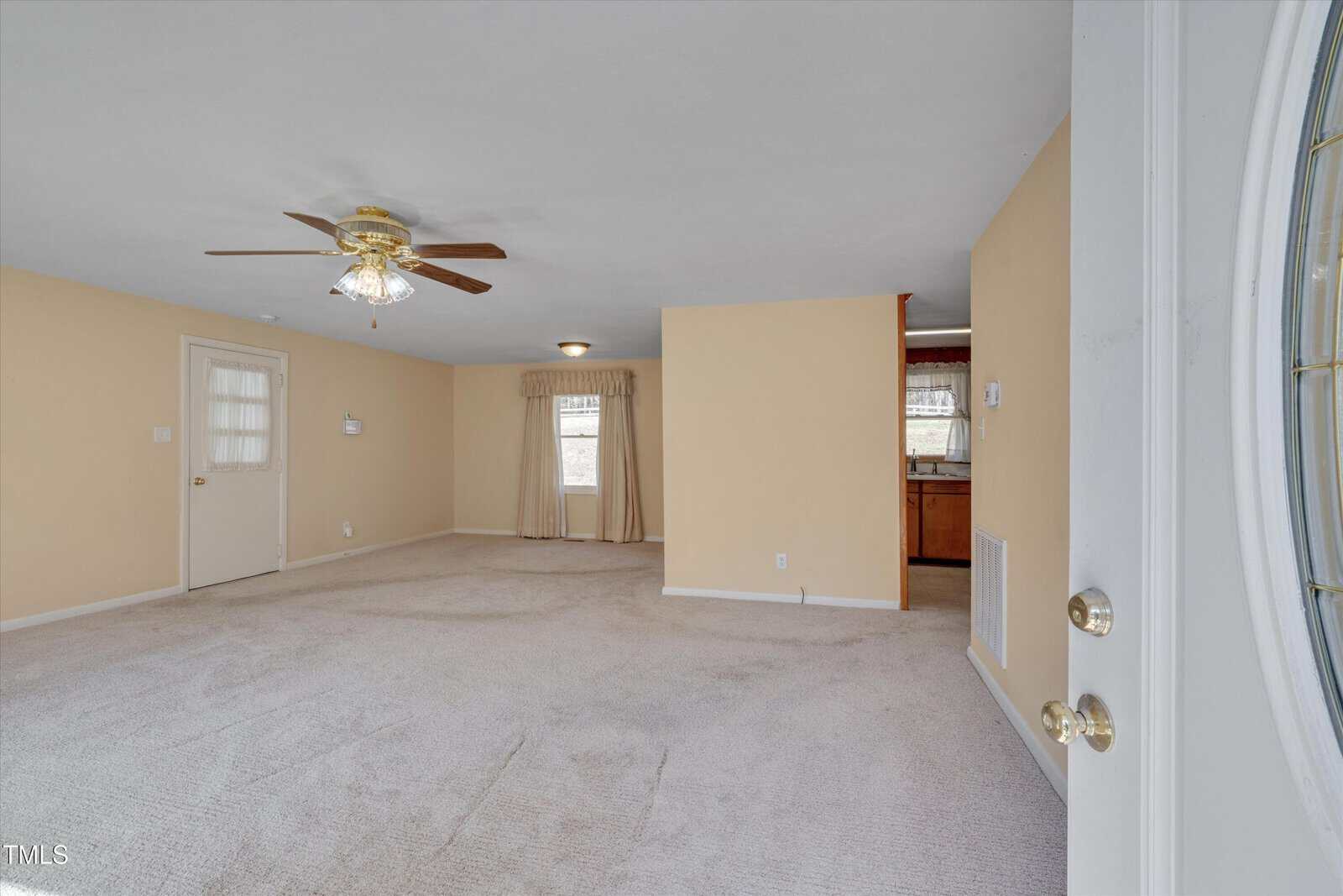 A spacious, carpeted living room with beige walls, a ceiling fan with a light, and a door leading outside. There is a window with curtains, and an open doorway reveals part of a kitchen with wooden cabinets.