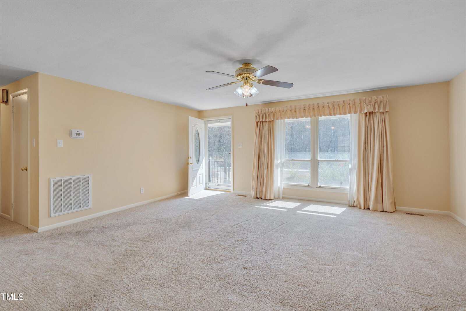 A spacious, carpeted living room with beige walls, a ceiling fan with lights, large front windows with curtains, an open door to the outside, and a vented wall panel on the left.